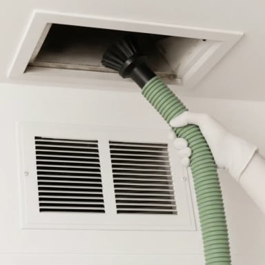 Hand using a vacuum hose to clean a ceiling vent.